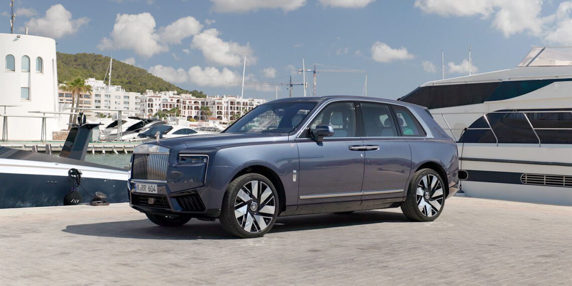 A luxury Rolls-Royce SUV, reflecting its record-breaking bespoke commissions, is elegantly parked beside yachts at a marina. Buildings rise nearby and a verdant hill stretches under the expansive blue sky dotted with clouds.