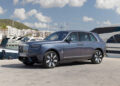 A luxury Rolls-Royce SUV, reflecting its record-breaking bespoke commissions, is elegantly parked beside yachts at a marina. Buildings rise nearby and a verdant hill stretches under the expansive blue sky dotted with clouds.