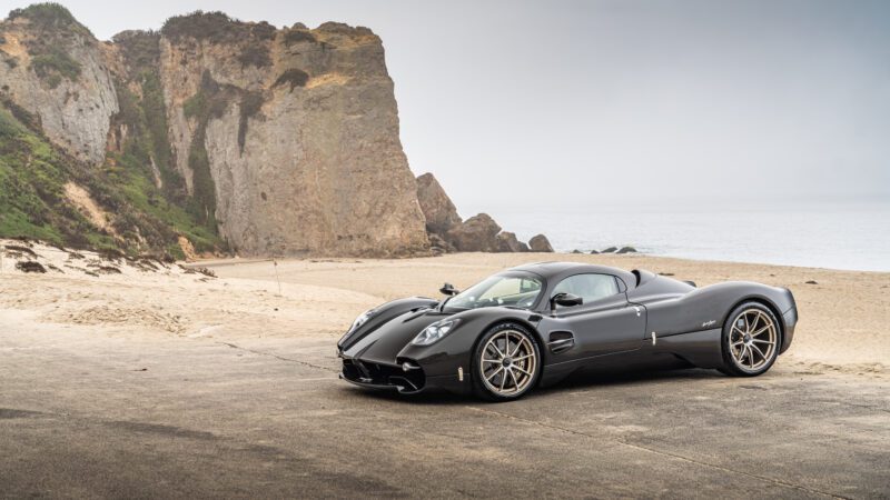 A sleek black sports car parked on a coastal road with rocky cliffs and ocean in the background.