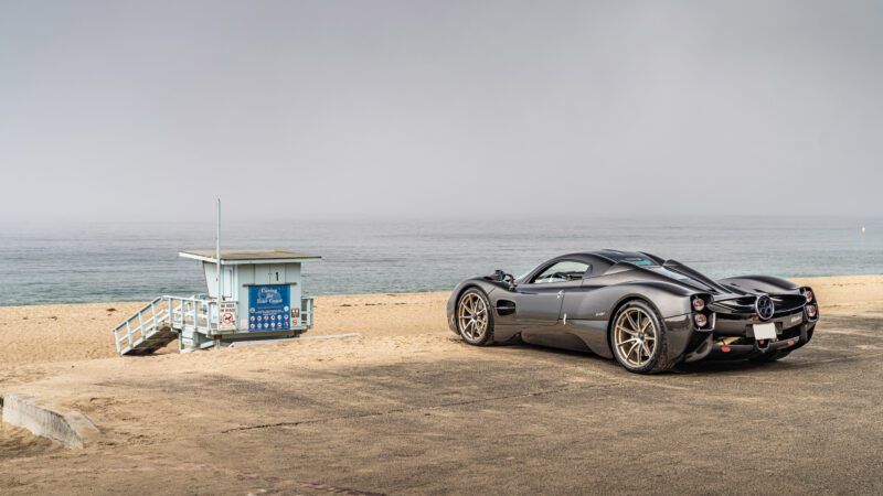 A sleek black sports car parked on a beachside road near a small blue lifeguard station with an overcast sky in the background.