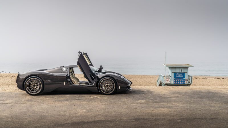 A sleek black sports car with open doors parked on a beachfront road next to a small lifeguard hut.