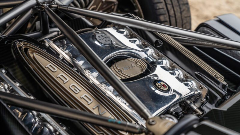 Close-up of a Pagani car's V12 engine, showing chrome details and structural elements, with a visible tire in the background.