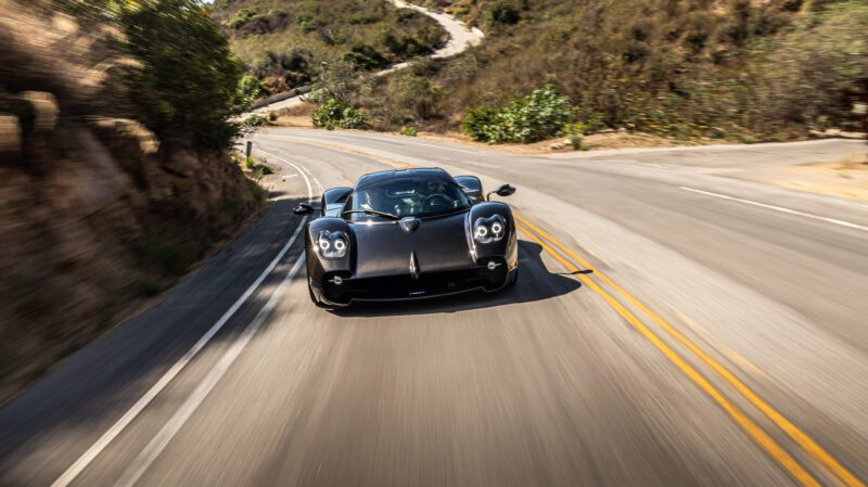 A black sports car speeds along a winding road surrounded by greenery and hills.