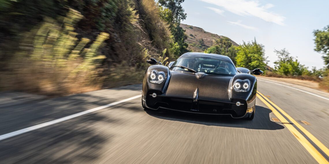 A sleek Pagani Utopia sports car glides along a scenic, winding road, embraced by lush greenery and rolling hills beneath a clear sky.