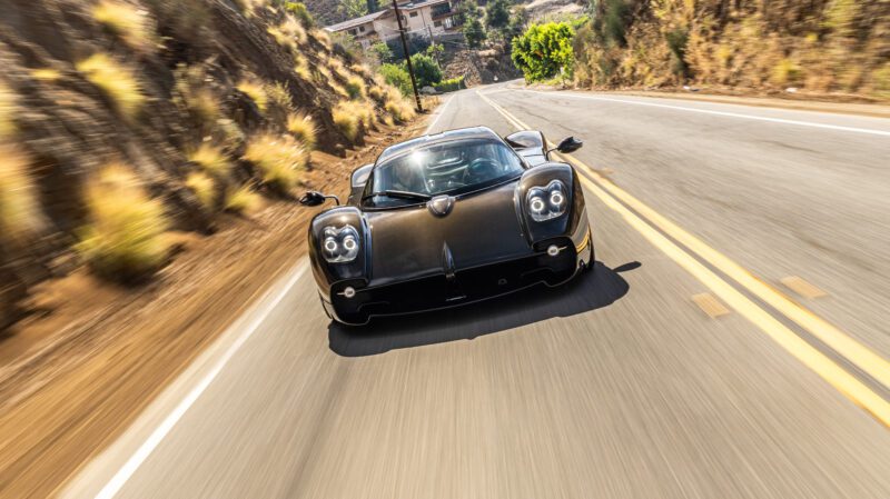 A black sports car speeds down a hilly road, surrounded by dry vegetation and trees.