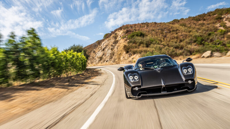 A sleek black sports car drives on a curved mountain road under a blue sky with clouds.