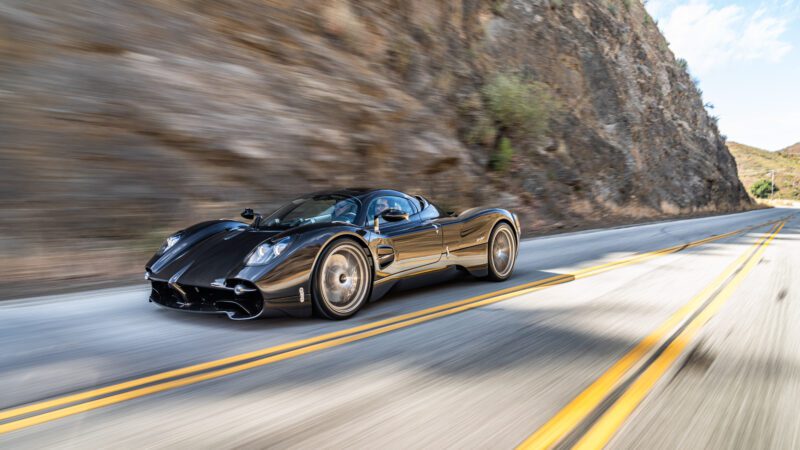 A sleek black sports car driving on a winding mountain road with rocky cliffs in the background.