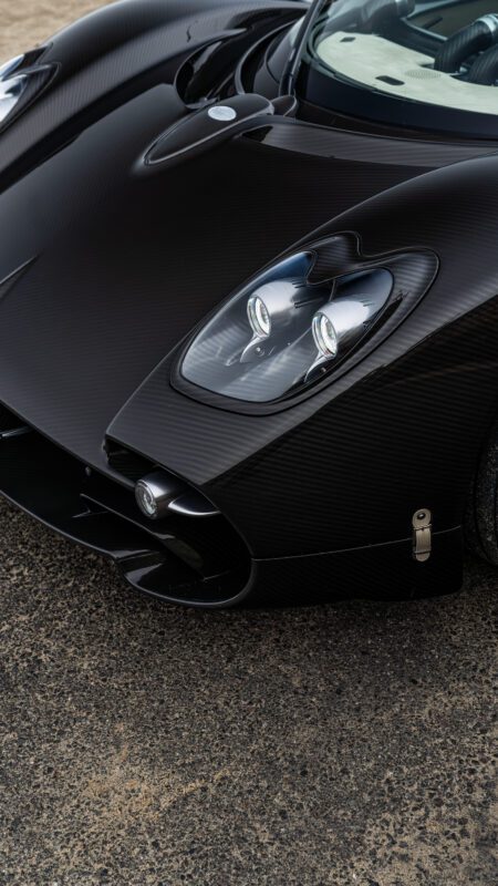 Close-up view of a sleek, black sports car front with distinct headlight design and carbon fiber finish, parked on a textured surface.