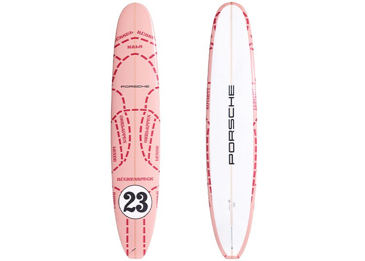 Two surfboards make waves in the 2024 Holiday Gift Guide: one pink with racing decals and a bold number 23, and another white featuring "Porsche" down the center, a standout suggestion from duPont REGISTRY.