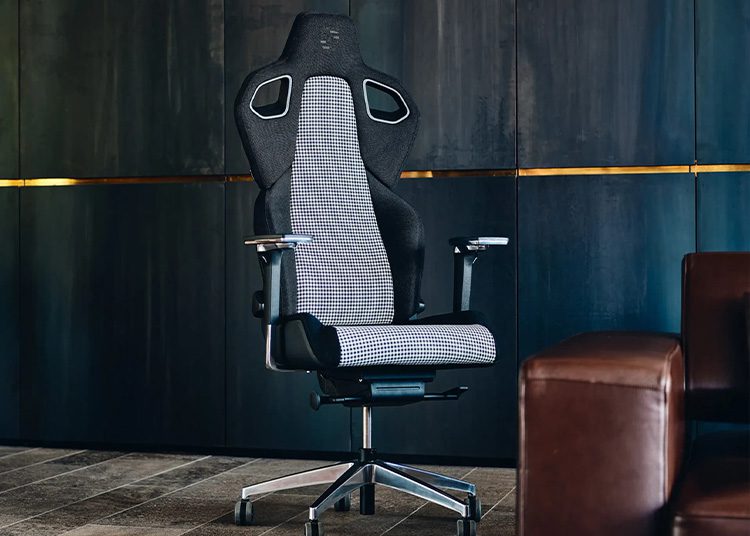 An ergonomic black and white office chair with armrests and a high back, featured in the 2024 duPont REGISTRY Holiday Gift Guide, is positioned on a tiled floor in front of a dark panel wall.