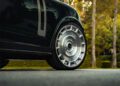 Close-up of a Custom-Built Urban Widetrack luxury car's front wheel, showcasing a detailed rim. It's parked on the road with the lush backdrop of trees and greenery offering a serene contrast to its bold design.