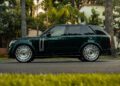 A sleek black Range Rover L460 with shiny rims is parked on a palm-lined street, its Custom-Built Urban Widetrack design turning heads amidst the backdrop of city buildings, reminiscent of Scott Disick's luxurious taste in vehicles.