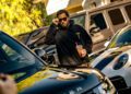 A person in a dark hoodie and sunglasses holds a drink beside a Custom-Built Range Rover, with other luxury cars visible in the background.