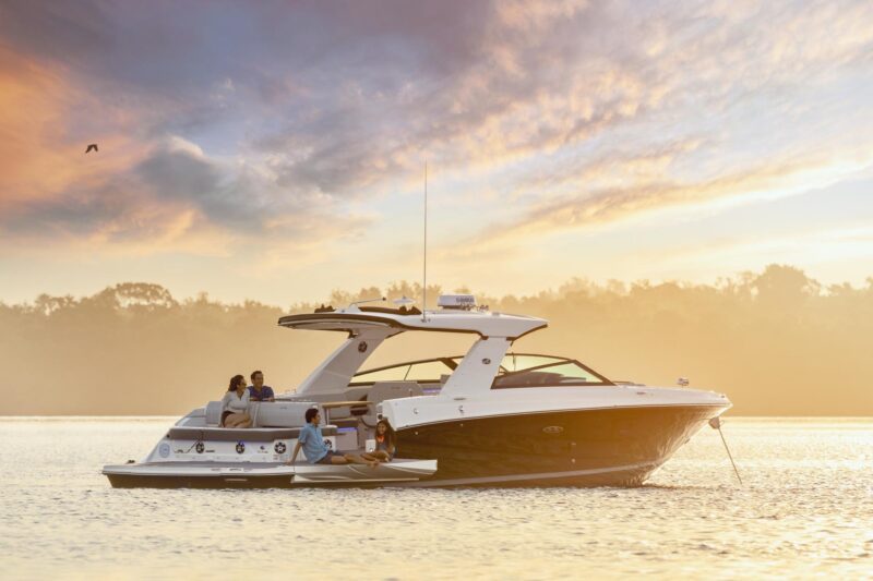 A group of people relax on a modern motorboat floating on calm water at sunset, enjoying a serene maritime experience against a backdrop of distant shoreline and cloudy sky.