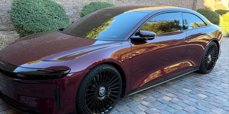 A sleek, custom Lucid Air in dark red with tinted windows is parked on a cobblestone driveway, set against a backdrop of brick walls and green shrubbery.