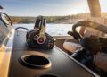 Close-up of a boat's steering console showcasing a throttle lever, cup holder, and steering wheel. This timeless design is set against the backdrop of water and distant shoreline under a clear sky, embodying luxury with every crafted detail.