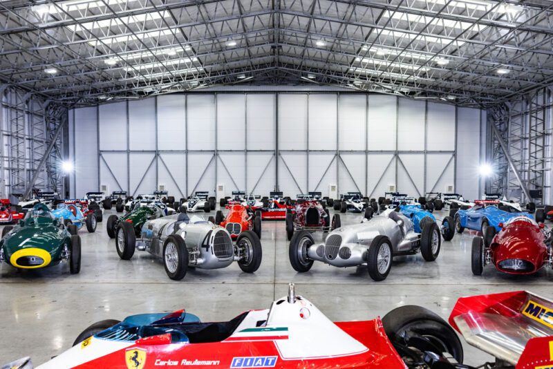 A vintage F1 car collection, once cherished by Bernie Ecclestone, is displayed in a spacious, well-lit hangar with a high metal ceiling.