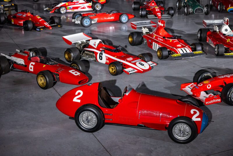 A captivating F1 car collection once admired by Bernie Ecclestone showcases various red vintage race cars on a gray concrete floor, each flaunting unique designs and numbers.