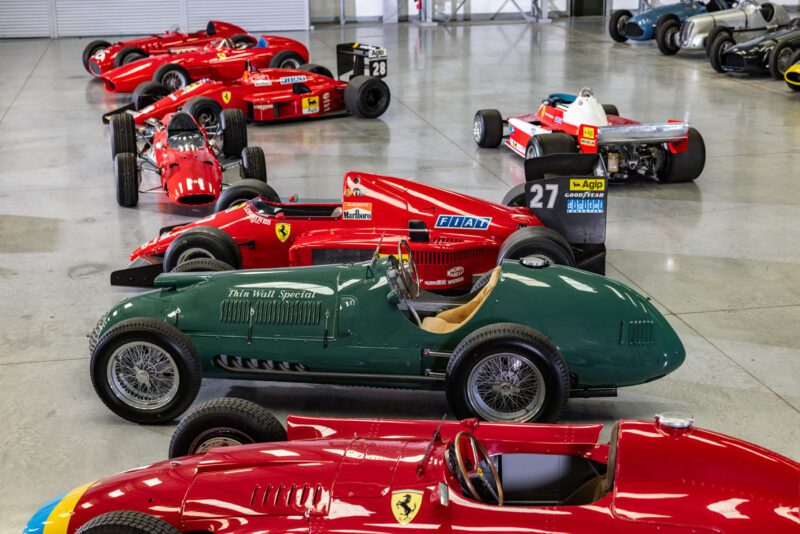 A collection of vintage and classic racing cars lined up in a showroom, featuring mostly red Ferraris and a green race car in the foreground. This exquisite display, once admired by Bernie Ecclestone, feels like an F1 Car Collection up for an exclusive sell.