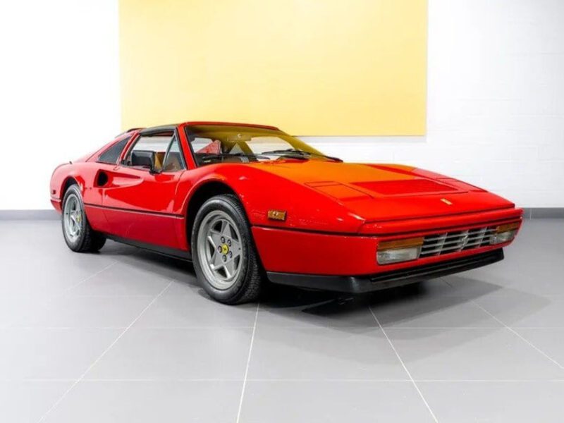 A red Ferrari 328 GTS sports car is parked indoors on a gray floor against a white wall with a yellow panel, symbolizing an exciting investment opportunity.