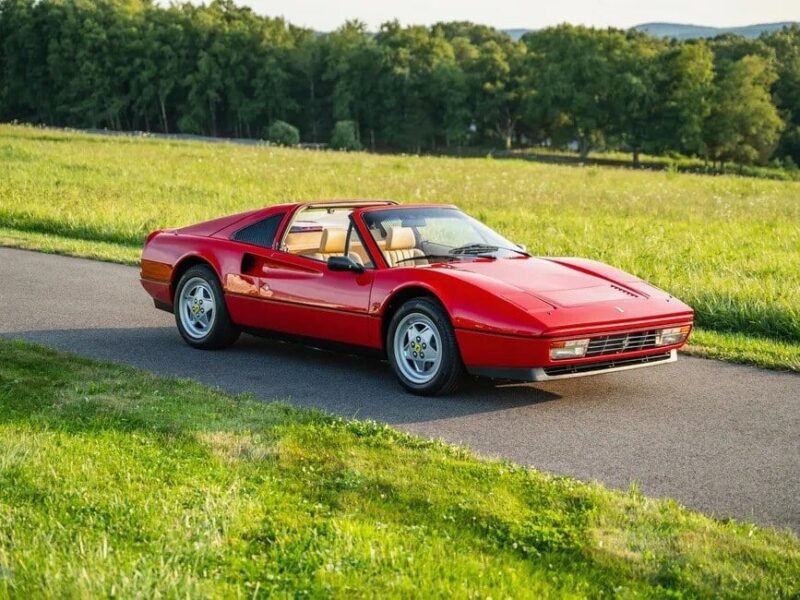 A Ferrari 328 GTS is parked on a narrow road in a grassy field with trees in the background, its sleek red frame embodying both elegance and precision.