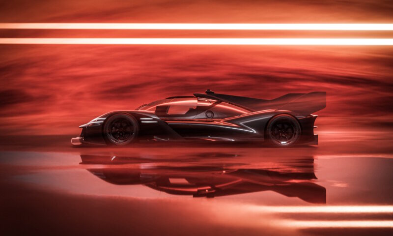 Side view of a sleek, futuristic race car with a prominent rear wing, echoing the spirit of Le Mans 24 Hours, set against a dramatic red-lit background with horizontal light streaks.