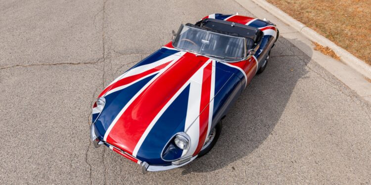 A classic Shaguar, boasting a Union Jack paint design, is parked on a paved road near a grassy area.
