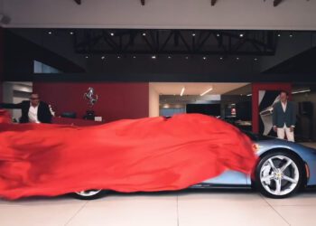A blue sports car being unveiled indoors with a red cloth being removed by three people.