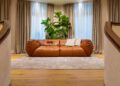 A brown leather sofa from the Home Collection, with two pillows, sits on a beige rug. Behind it is a large green plant and windows with beige curtains, adding a touch of Parisian elegance.