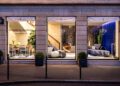 Storefront with three large windows showcasing the Bugatti Home Collection, featuring luxury home furnishings, including seating and decor. The display is illuminated with soft lighting, and two potted plants are outside, evoking the elegance of Parisian style.