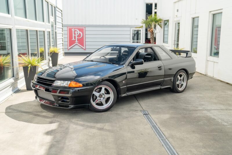 A black Nissan Skyline GT-R R32, a true icon of 90s cars for sale, is parked on a concrete driveway outside a modern building with glass windows and potted plants.