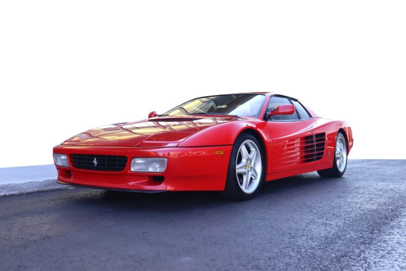 A red Ferrari sports car, evoking nostalgia for classic cars, is parked on a paved surface with a white background, viewed from the front-left angle.
