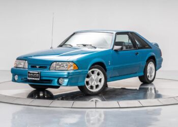 A blue Ford Mustang coupe, reminiscent of 90s cars, is displayed on a turntable in a studio with a plain white background, evoking a sense of nostalgia.