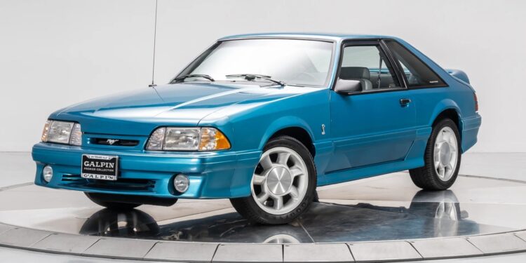 A blue Ford Mustang coupe, reminiscent of 90s cars, is displayed on a turntable in a studio with a plain white background, evoking a sense of nostalgia.