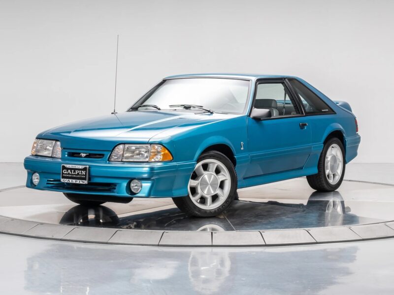 A blue Ford Mustang coupe, reminiscent of 90s cars, is displayed on a turntable in a studio with a plain white background, evoking a sense of nostalgia.