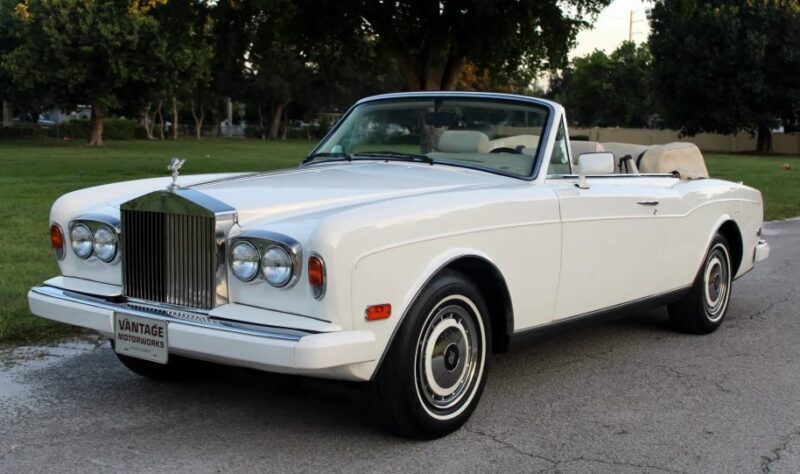 A white vintage convertible luxury car is parked on a paved road, framed by a grassy area and trees in the background.