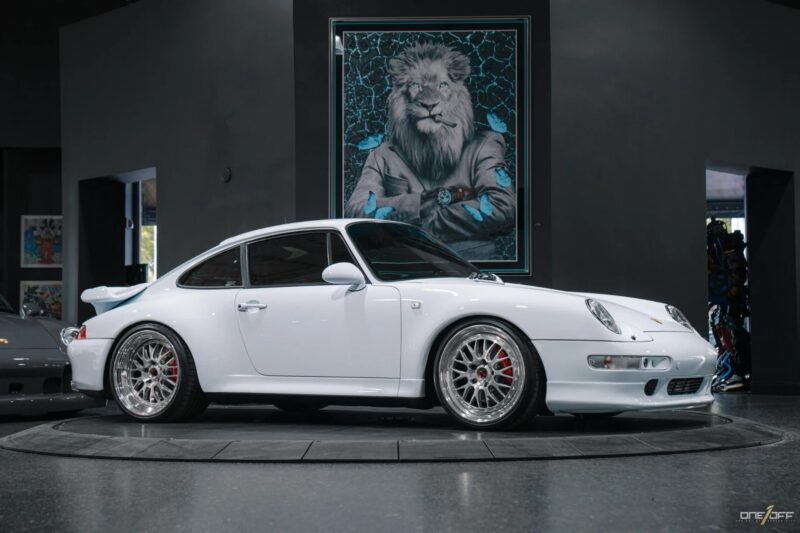 A white Porsche 911 sports car, a true icon of 90s cars for sale, is parked indoors in front of a framed artwork depicting a lion in a suit with folded arms.
