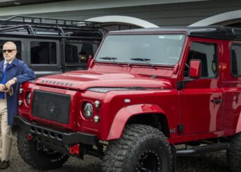 A man in a blue coat and sunglasses stands next to a red off-road vehicle, highlighting the Helderburg Defender Restorations legacy, with a dog sitting nearby. Another similar vehicle, a testament to modern luxury, looms in the background.