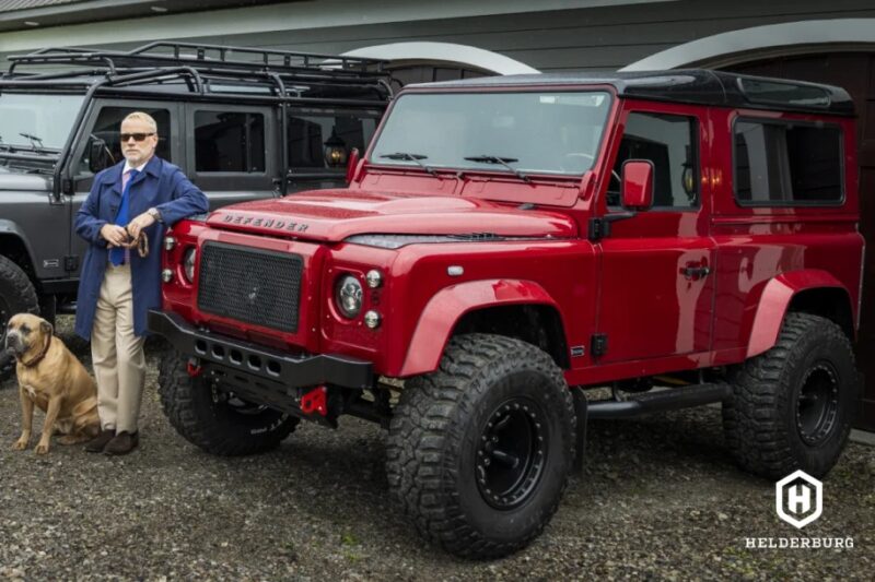 Seseorang dengan mantel biru berdiri dengan seekor anjing besar di sebelah kendaraan 4x4 merah, menunjukkan hasrat mereka untuk di luar jalan, dengan 4x4 hitam lain yang diparkir di belakang mereka.