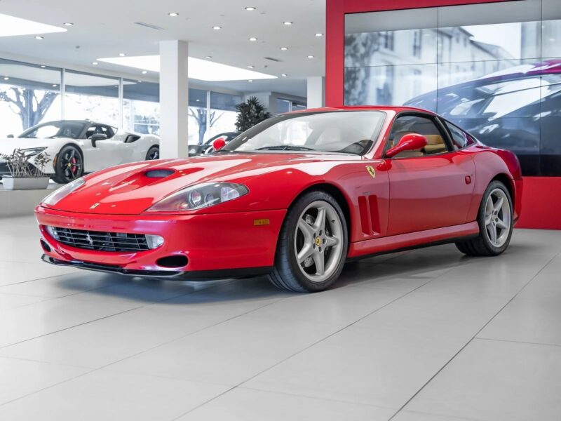 A red Ferrari sports car is parked indoors in a modern showroom with large windows, surrounded by other cars that evoke a sense of nostalgia and history.