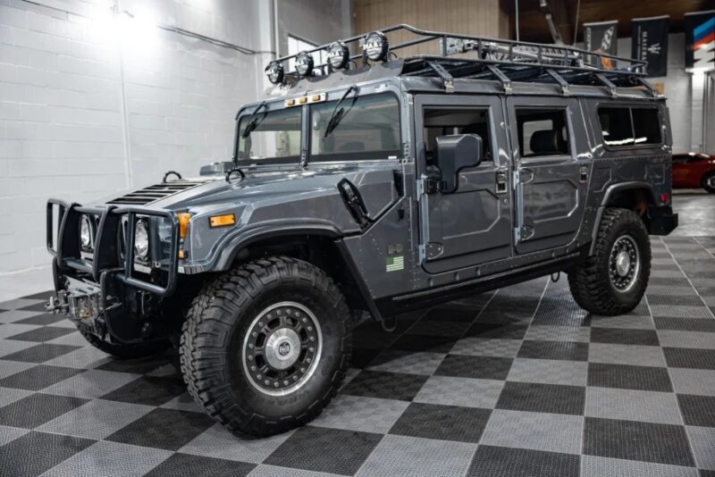 A gray Hummer H1, an exotic car adorned with off-road lights and a roof rack, stands proudly indoors on a checkered floor. Ideal for adventure enthusiasts, this rugged beauty is now for sale, offering both power and prestige to its next owner.