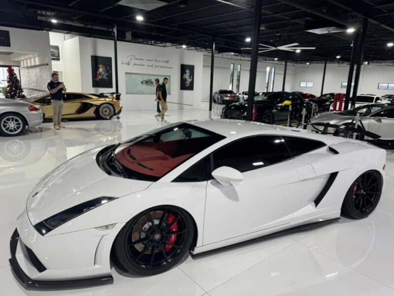 A striking white Lamborghini Gallardo with black wheels is showcased indoors among other luxury cars. This V10 supercar draws attention as two people walk by in the background, contemplating if this masterpiece is for sale.