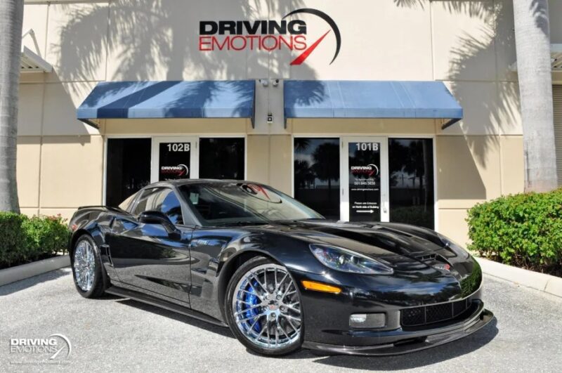 An exotic car with blue and silver alloy wheels is parked in front of a building adorned with a "DRIVING EMOTIONS" sign, surrounded by swaying palm trees. This luxury sports car offers an enticing opportunity for those seeking high-performance elegance.