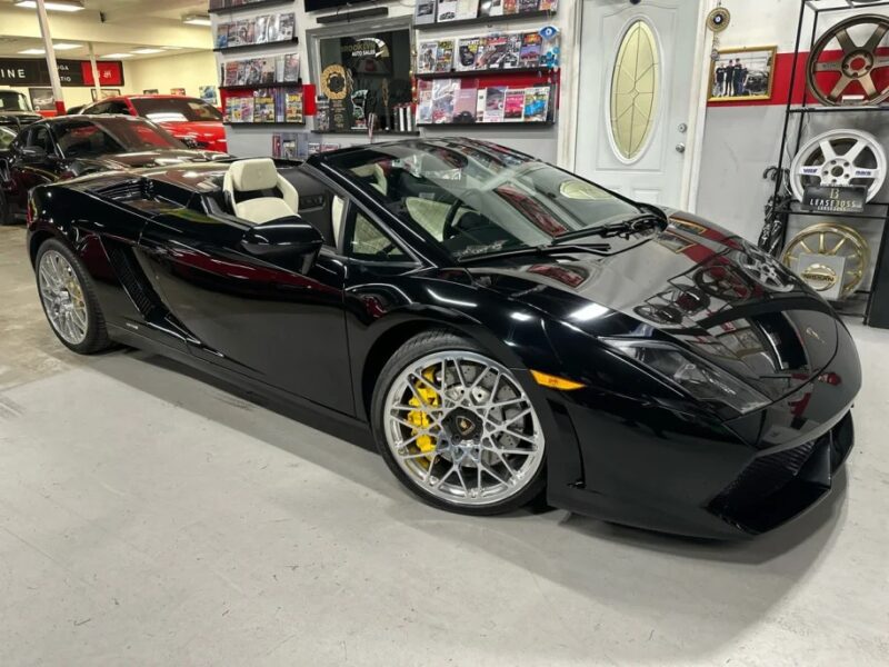 A stunning black convertible sports car with yellow brake calipers is parked indoors, surrounded by various automotive memorabilia on the walls. This sleek V10 supercar is reminiscent of Lamborghini Gallardos and could be your dream car for sale.