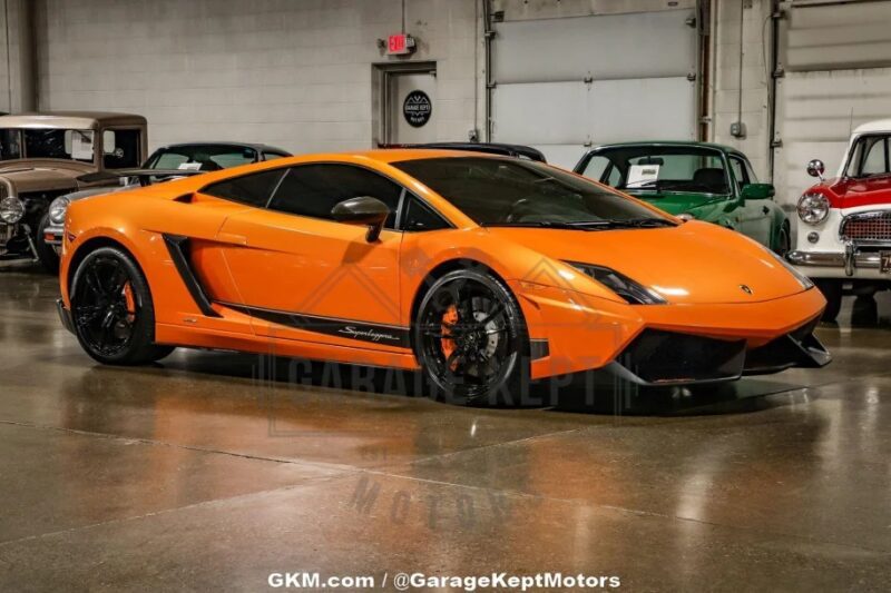 A striking orange V10 supercar sits parked indoors, with classic cars in the background. This sleek Lamborghini Gallardo is now for sale, offering a rare opportunity to own a piece of automotive excellence.