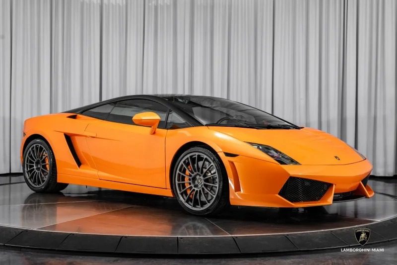 A striking orange Lamborghini Gallardo V10 supercar is elegantly displayed in the showroom, set against a sleek gray curtain background, and is now available for sale.