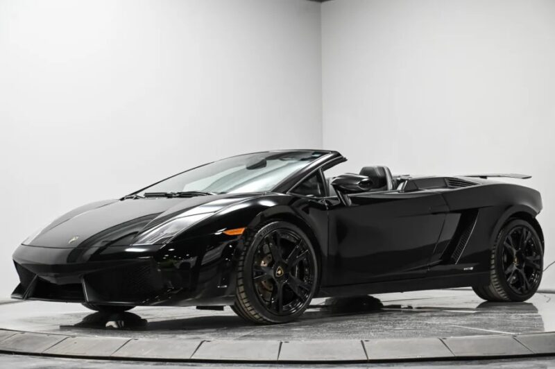 A black convertible sports car, possibly one of the coveted Lamborghini Gallardos, with its top down, is parked in a well-lit indoor space. The V10 supercar flaunts its sleek design from a front three-quarter angle.