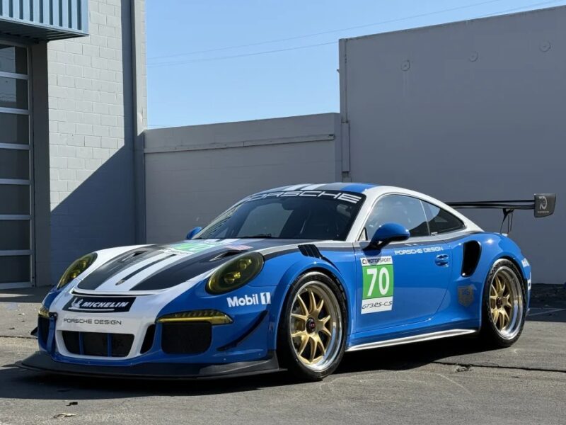 The blue and white Porsche race car, numbered 70 and adorned with sponsorship logos, is an exotic luxury car parked on a paved surface in front of a gray building.