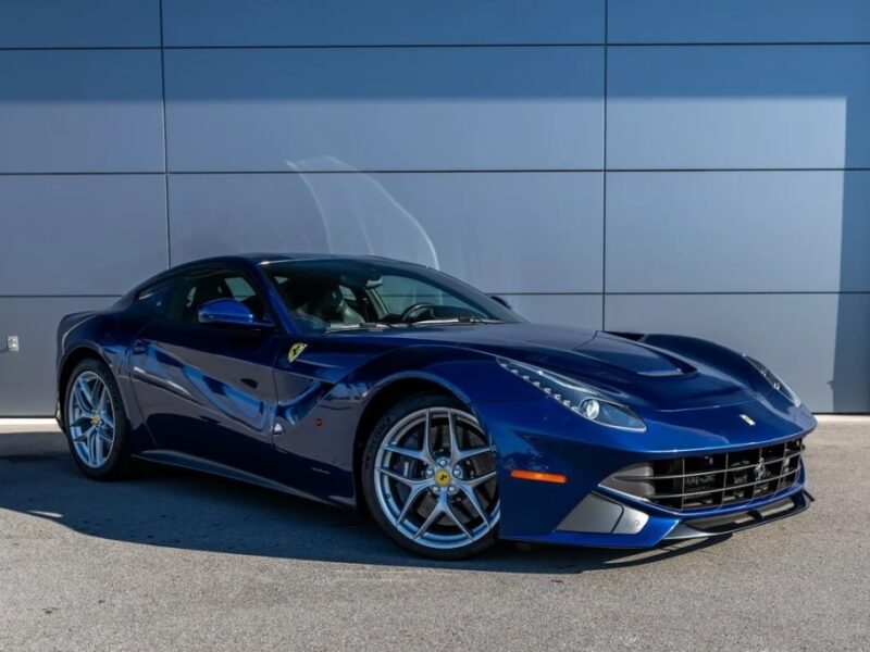 A blue Ferrari sports car, an epitome of exotic luxury, is parked on a paved surface in front of a gray wall, viewed from the front corner.