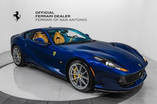 An exotic blue Ferrari sports car with yellow brake calipers and a tan interior is showcased in the showroom. Under Ferrari of San Antonio branding, this luxury car for sale stands as a testament to elegance and performance.